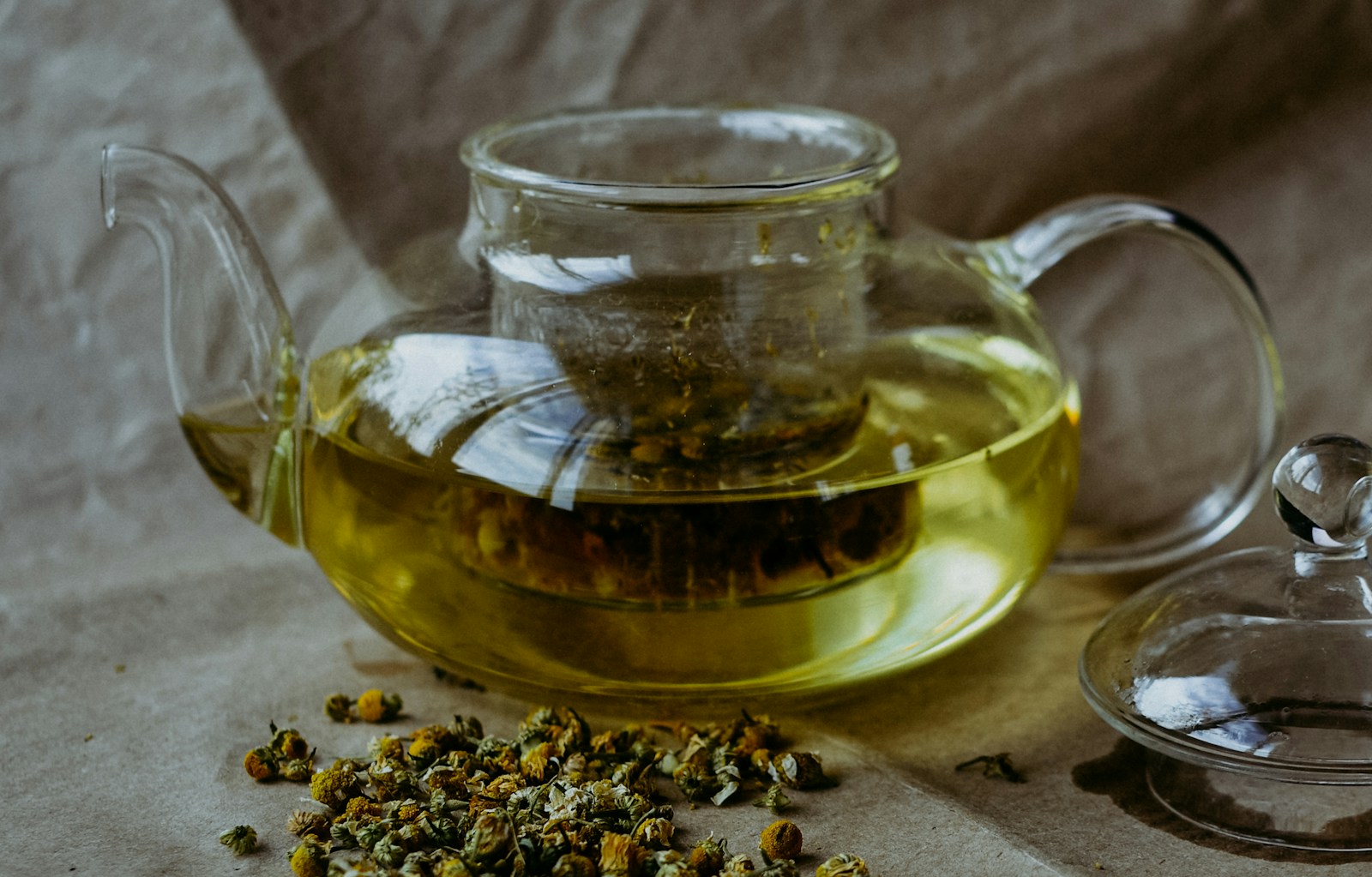Glass teapot of chamomile tea beside a pile of dried chamomile flowers