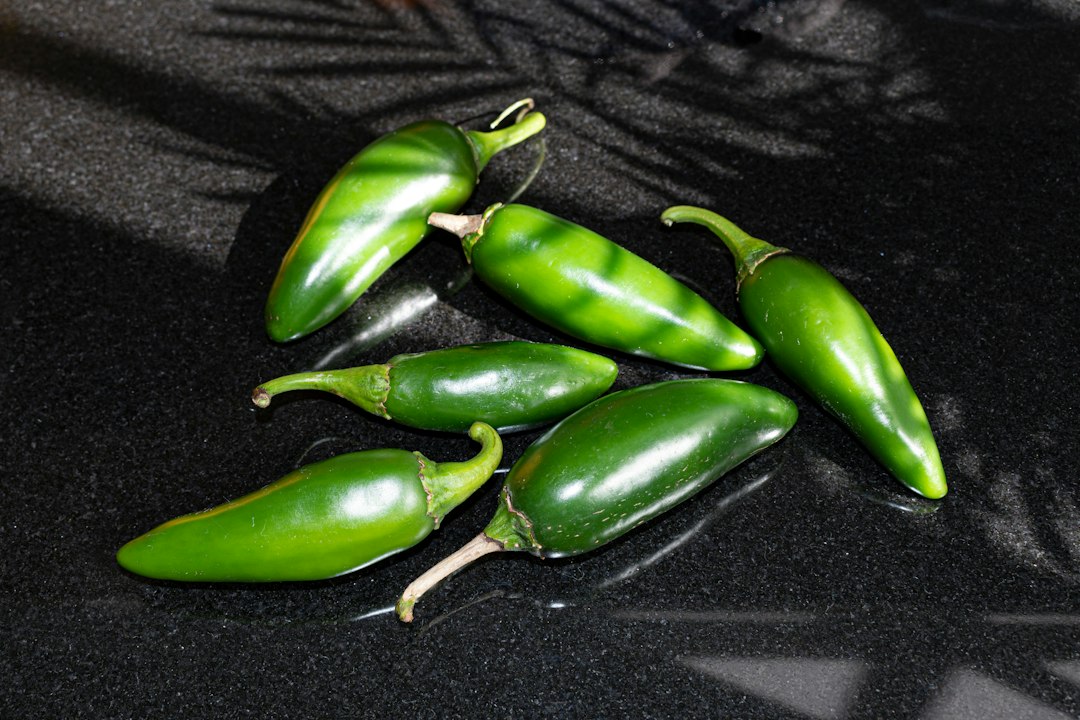 Six glossy green jalapeño peppers with curled stems on a dark counter