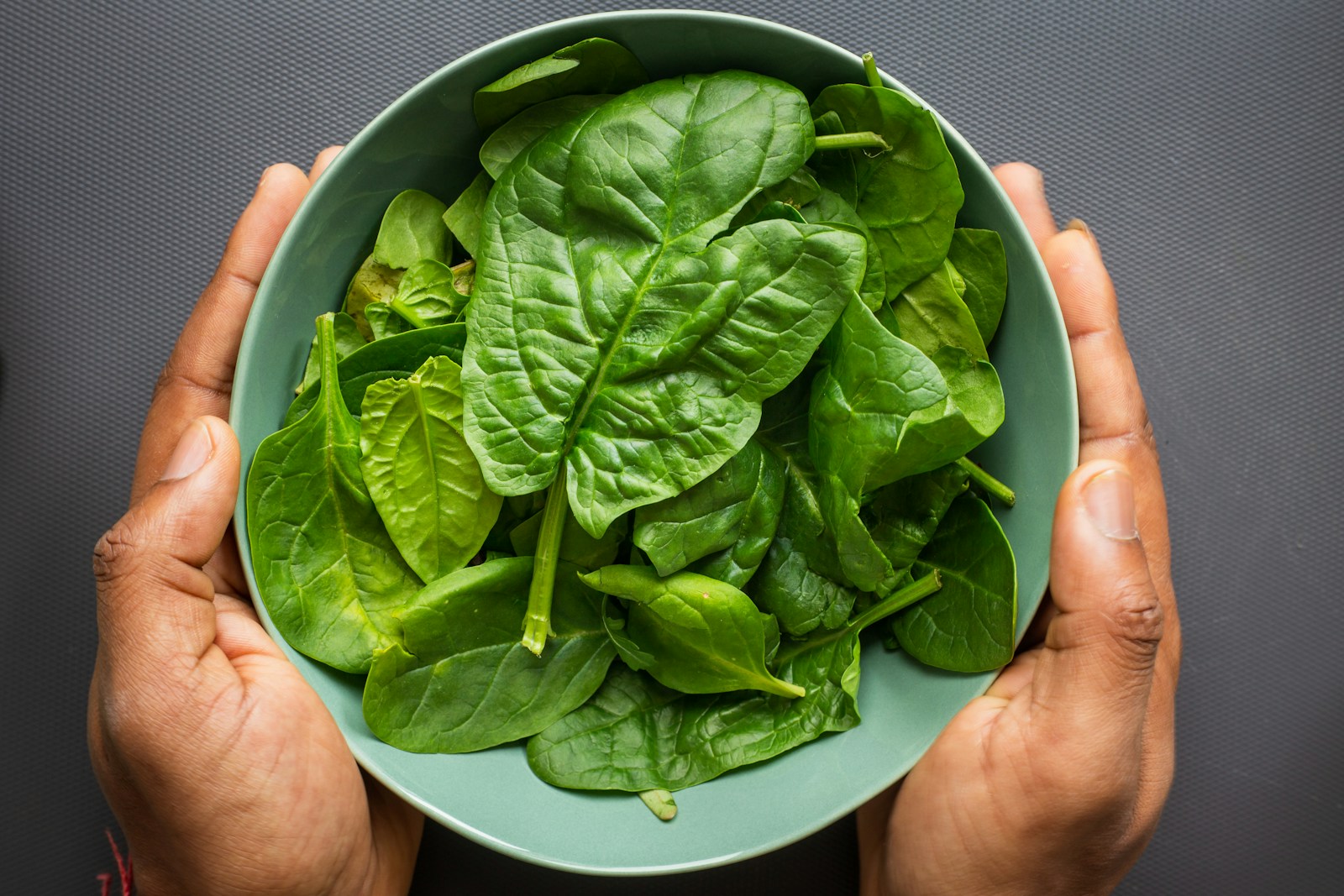 Two hands holding a green bowl filled with fresh spinach leaves
