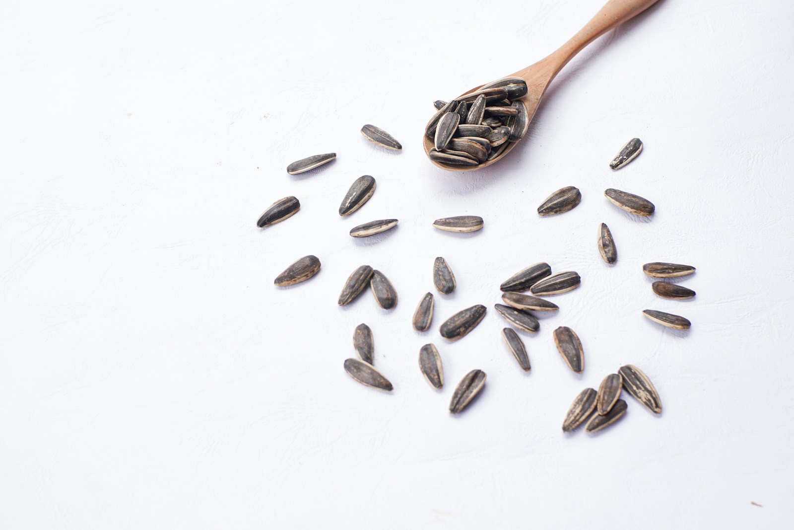 Grey-striped sunflower seeds scattered around a wooden spoon on white